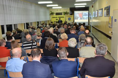 W szkolnej klasie trwa spotkanie z mieszkańcami. Szkolna sala jest wąska, po lewej okna. Zdjęcie zrobione z tyłu klasy, zza pleców ludzi siedzących na krzesłach. Na przodzie sali zwrócony do ludzi stoi prezydent miasta. Obok po prawej ekran, na którym jest wyświetlana prezentacja. Po lewej baner z napisem: Zarząd Osiedla 