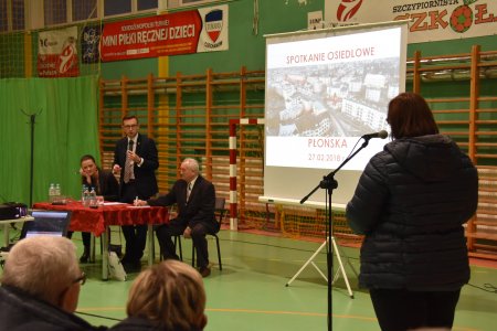 Spotkanie z mieszkańcami w sali gimnastycznej (drabinki gimnastyczne na ścianach). Na pierwszym planie kobieta mówi do mikrofonu zamocowanego na statywie. Na drugim planie za stolikiem, przy którym siedzą przedstawiciele Urzędu Miasta, stoi prezydent Ciechanowa.