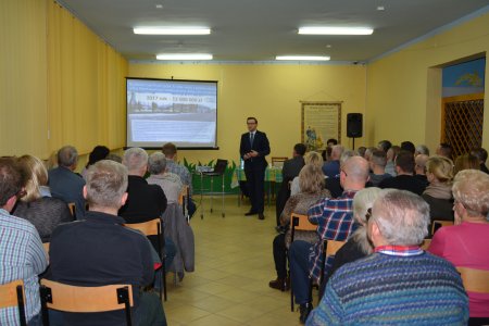 Spotkanie z prezydentem. Zdjęcie zza pleców mieszkańców, naprzeciw których stoi prezydent. Obok elran z wyświetlaną prezentacją