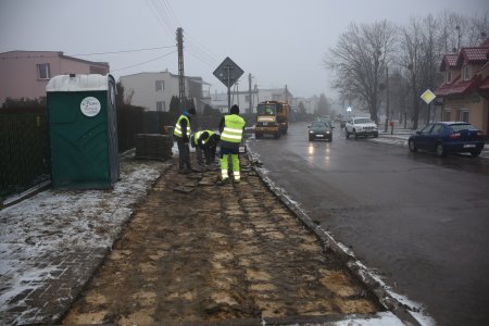 Po lewej trzech robotników w odblaskowych kamizelkach, wymaganych podczas prac na drodze, zdejmują stare, betonowe płyty z chodnika. Po prawej jezdnia i jeżdżące nią samochody