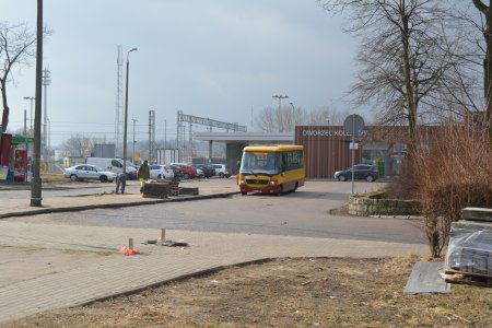 Plac przed dworcem kolejowym. Widać parkujące tu samochody i autobus stojący na przystanku