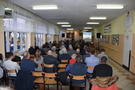 W szkolnej sali trwa spotkanie z mieszkańcami. Zdjęcie wykonane zza pleców mieszkańców siedzących na krzesłach. W oddali stoi ekran, na którym wyświetlana jest prezentacja