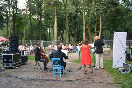 Koncert na wolnym powietrzu. Na pierwszym planie grający muzycy. W głębi siedzi publiczność