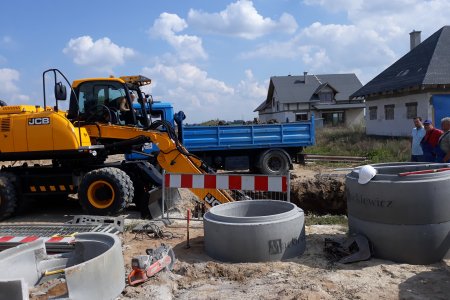 Koparka wykonuje głęboki wykop. W pobliżu leżą duże betonowe kręgi czekające na ułożenie w wykopie