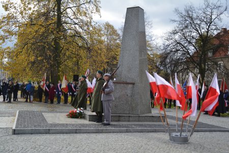 Przy pomniku marszałka Józefa Piłsudskiego trwa uroczystość. Przy pomniku stoi czteroosobowa warta służb mundurowych. Po obu stronach ustawiono stojaki z kilkoma narodowymi flagami każdy. Za pomnikiem stoją uczestnicy uroczystości. Za nimi widać drzewa ze skweru im. marszałka