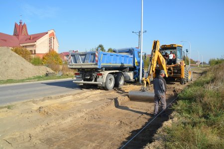 Koparka z szeroką łyżką zbiera wierzchnią warstwę ziemi w miejscu budowy chodnika. Ziemię wysypuje na stojącą obok wywrotkę. W oddali widać fragment kościoła św. Franciszka z Asyżu