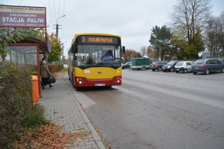 Autobus miejskiej linii nr 3 stoi na przystanku naprzeciwko cmentarza komunalnego.  Dwie osoby podchodzą do autobusu. Na dachu przystanku reklama stacji paliw prowadzonej przez Zakład Komunikacji Miejskiej.