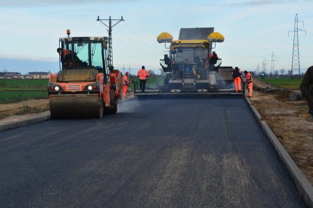 Duże drogowe maszyny równają nawierzchnię jezdni. Pierwszy jedzie wale równający część pasa drogowego. Za nim maszyna obejmująca całą szerokość jezdni. Przy tej większej maszynie jest kilku robotników. Po lewej stronie pola, a za nimi domy mieszkalne. Po prawej na polach rząd słupów wysokiego napięcia