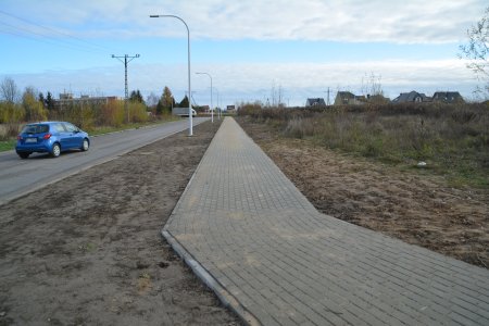 Pośrodku zdjęcia (pionowo) biegnie równiutki chodnik z brukowej kostki. Po lewej fragment biegnącej po skosie jezdni, po której jedzie samochód. Po prawej fragment gruntu porośniętego trawą