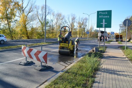 Za ustawioną na jezdni barierką trwa wyrównywanie asfaltu. Pracuje tam nieduży walec drogowy. Robotnik przy pomocy łopaty przygotowuje miejsce, które ma wyrównać walec. Na pasie zieleni między jezdnią i chodnikiem stoi drogowskaz 