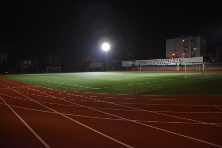 Zdjęcie wykonane po zmroku. Na wprost światło lampy oświetlającej boisko - okrągły jasny punkt i fragment jasnej murawy. Na dole zdjęcia bieżnia z liniami wyznaczającymi tory dla biegaczy. Na tyłach boiska ściana z muralem: Ciechanów 100% LEGIA