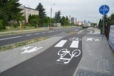 Wyremontowana ulica. Od lewej budynek wśród drzew, chodnik, wąski pas zieleni, jezdnia, znów wąski pas zieleni, chodnik, ścieżka rowerowa i znów chodnik. Na ścieżce rowerowej przejście dla pieszych. W oddali jadące auto