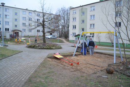 Trwa ustawianie urządzeń na placu przy blokach mieszkalnych. Alejki między blokami prowadzą do okrągłej wysepki porośniętej trawą, na środku której rośnie drzewo. Woków tego okrągłego, wypukłego trawnika wiedzie także okrągły chodnik