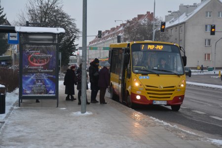 Przystanek autobusowy przy placu Jana Pawła II. Na przystanek podjechał miejski autobus. Pasażerowie w drodze do drzwi pojazdu. Na przystankowej szybie wisi duży plakat zapowiadający koncert noworoczny 