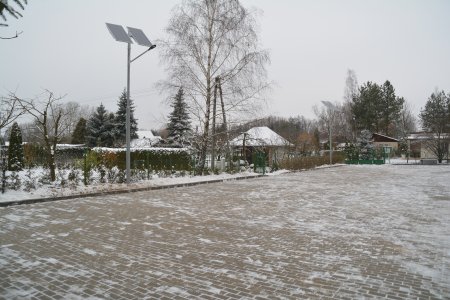 Zima. Spory plac wyłożony betonową kostką. Od góry chodnik, z za nim ogrodzenie i żywopłot. Pośrodku chodnika lampa z własnym zasilaniem na baterie słoneczne