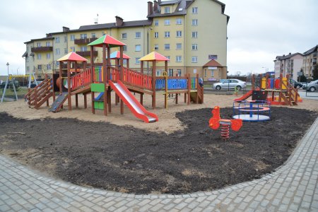 Plac zabaw, a w nim karuzela, kilka daszków i przejść między nimi, zjeżdżalnia, bujak. Na pierwszym planie miejsce na trawnik, dalej plac wysypany żwirem