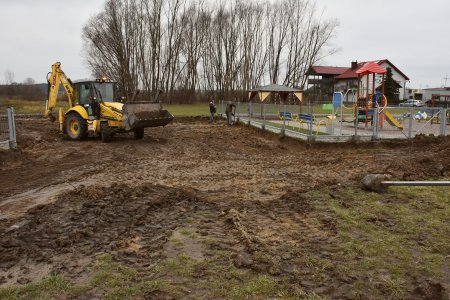 Po lewej teren rozjeżdżony przez ciężki sprzęt. Jeździ tu koparka. Po prawej ogrodzony plac zabaw. W oddaliwidać wysokie drzewa i dom