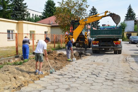 Trwa remont drogi wyłożonej betonowymi sześciennymi elementami. Elementy te zdejmuje koparka, która ładuje je na samochód z przyczepą. Na części pozbawionej betonowych bloków pracują robotnicy drogowi, którzy wyrównują teren