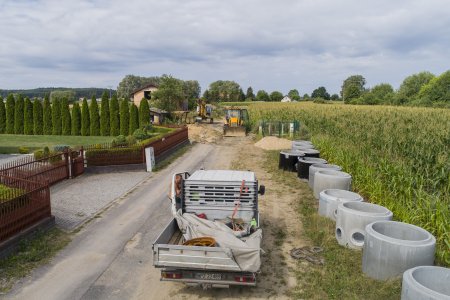 Gruntowa droga biegnąca między prywatną posesją z lewej i polem kukurydzy z prawej. Wzdłuż drogi ułożony jest rząd betonowych kręgów. Na drodze stoi ciężarówka, a dalej inne maszyny