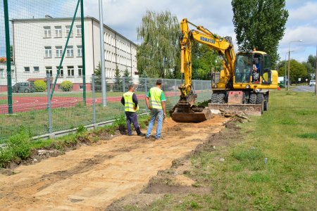 Wzdłuż ogrodzenia boiska przy szkole podstawowej nr 6 koparka zrywa trawę. Przygotowuje miejsce pod budowę drogi dla pieszych i rowerzystów. Przed koparką stoi dwóch robotników drogowych w ochronnych kamizelkach