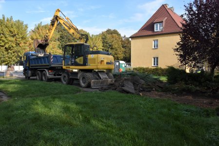 Wielka koparka zrywa nawiechchnię starego chodnika, który biegnie obok terenu porośniętego trawą. W tle budynek wielorodzinny, typowy dla zabudowy osiedla Bloki. Wokół rosną wysokie gęste drzewa