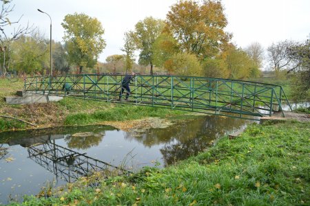 Kładka nad rzeką. Zbudowana z metakowych prętów. Na kładce stoi mężczyzna. Kładka odbija się na powierzchni wody. Po obu brzegach rzeki rośnie trawa. W oddali drzewa