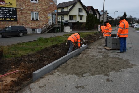 Kilku pracowników w odblaskowych kamizelkach układa krawężnik wzdłuż betonowej nawierzchni. Z połowy trawnika po lewej zebrano warstwę ziemi. W głębi kadru widać zabudowania