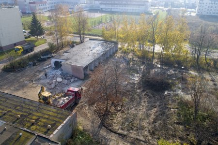 Zdjęcie wykonane z wysoka. Teren zaniedbany, nierówna zabłocona nawierzchnia. Po lewej budynek w trakcie rozbiórki. Obok betonowy gruz. Niedaleko budynku stoi koparka i samochód ciężarowy