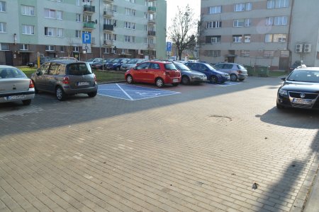 Wyłożony brukową kostką parking między blokami. Zaparkowane auta. Wolne miejsce zarezerwowane dla osób niepełnosprawnych