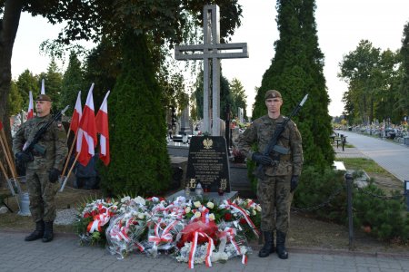 Na miejskim cmentarzu po obu stronach krzyża o wysokości około 3m stoją dwaj żołnierze. W dolnej części krzyża tablica z orłem na górze i napisami poniżej. Przed krzyżem leżą wiązanki kwiatów. Za plecami żołnierza po lewej widać stojak, w którym ustawiono narodowe flagi.