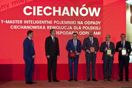 Siedmiu samorządowców stoi na tle dużego, czerwonego bajeru z napisem: Ciechanów T-master inteligentne pojemniki na odpady, ciechanowska rewolucja dla polskiej gospodarki odpadami. Pośród samorządowców stoi prezydent Ciechanowa i prezentuje nagrodę