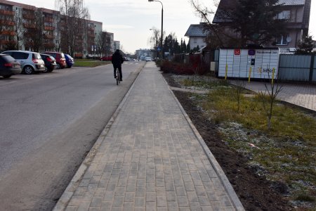 Pośrodku zdjęcia nowy chodnik wzdłuż jezdni. Od lewej widać blok, parking z autami i jezdnię.Po prawej stronie chodnika trawnik i dalej paczkomat