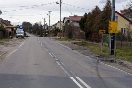 Ulica Gruduska ze zniszczoną nawierzchnią i piaszczystymi poboczami bez chodników i ścieżki dla rowerów. Po obu stronach ulicy prywatne posesje z domami jednorodzinnymi. Po lewej stronie w oddali widać przystanek autobusowy ze słupkiem i małym zadaszeniem.