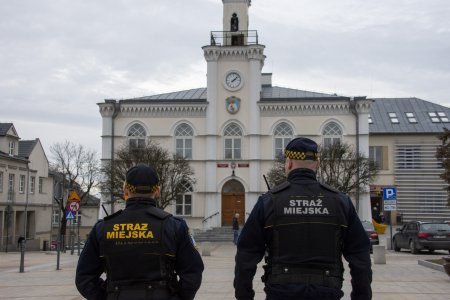 Dwóch strażników miejskich stojących na placu Jana Pawła 2. Stoją tyłem do fotografującego i przodem do ratusza. Na plecach mundurów mają napis: straż miejska.