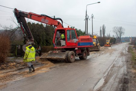 Wąska asfaltowa ulica. Po obu stronach błoto. Po lewej stronie stoi koparka, która zbiera wierzchnią warstwę gruntu wzdłuż asfaltu. Obok koparki stoi robotnik w odblaskowej kurtce i gumowcach. Sporo dalej, po tej samej stronie drogi stoi spycharka