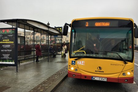 Miejski autobus linii 3 stoi na przystanku. Po lewej przystankowa wiata, a na niej plakat informujący o Jarmarku Bożonarodzeniowym. Na przystanku stoi kilka osób