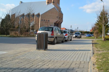 Chodnik po prawej stronie ul. Kraszewskiego biegnącej do kościoła św. Piotra. W głębi widac kościół. Wzdłuż chodnika, na jezdni parkuje rząd aut