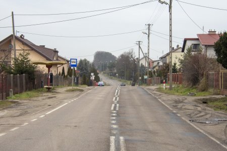 Ulica ze zniszczoną nawierzchnią. Podzielona liniami na dwa pasy przeciwnych kierunków ruchu. Widoczne linie brzegowe jezdni. Po obu stronach nierówne, gruntowe pobocze. Brak chodników. Po lewej stronie widać zadaszony przystanek autobusowy i niedużym siedzeniem dla pasażerów. Po obu stronach ulicy domy jednorodzinne