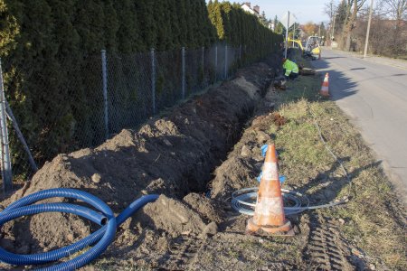 Wzdłuż ulicy po jej lewej stronie wykopano rów, w którym ułożono już niebieską rurę z tworzywa o średnicy kilku centymetrów. Nadmiar rury wystaje zwinięty na bliższym końcu rowy. Obok rowy leży wąska, biała taśma. Wzdłuż rowy od strony ulicy stoi kilka słupków w paski. W oddali widac pracownika, który kuca przy rowie