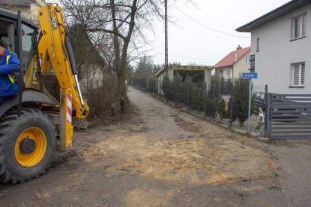 Wąziutka dróżka między posesjami o nawierzchni gruntowej. Po lewej stoi koparka. Przy ogrodzeniu posesji po prawej stronie na słupku przymocowana jest tabliczka z nazwą 