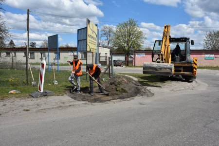 Na poboczu drogi dwóch pracowników drogowych łopatami zdejmuje wierzchnią warstwę gruntu. Powstanie tutaj chodnik. W pobliżu stoi koparka.