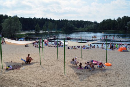 Plaża na Krubinie. Na pierwszym planie znajdują się dwa żagle przeciwsłoneczne. To duże, białe, trójkątne kawałki tkaniny, rozwieszonej na trzech wysokich i cienkich słupach. Tkanina daje cień, w którym można się schować przed palącym słońcem. Pod żaglami siedzi kilka osób. W głębi znajduje się dalsza część plaży i kąpielisko z pomostem. W głębi zdjęcia widać drzewa, rosnące wokół zbiornika wodnego.