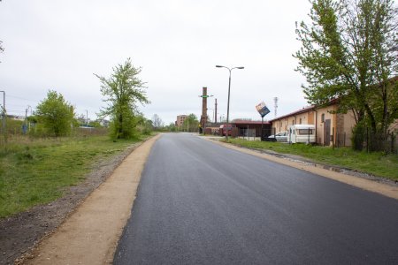 Na zdjęciu znajduje się fragment ulicy z ciemną, równiutką nawierzchnią. Wzdłuż drogi ciągną się wyrównane, żwirowe paski pobocza. Po lewej stronie ulicy jest trawnik i kilka krzewów. Po prawej, za ogrodzeniem znajduje się podłużny budynek Hurtowni Sigma, przy którym stoi biała przyczepka i auto. W oddali widać komin ciechanowskiej cukrowni.