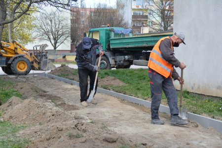 Dwóch pracowników przygotowuje podłoże pod chodnik. Z jednej strony ułożony już został krawężnik, z drugiej rozciągnięto białą linkę w miejscu, gdzie będzie drugi krawężnik. Po prawej stronie zdjęcia widać fragment bloku, a w tle widać niedużą koparkę i zieloną wywrotkę.