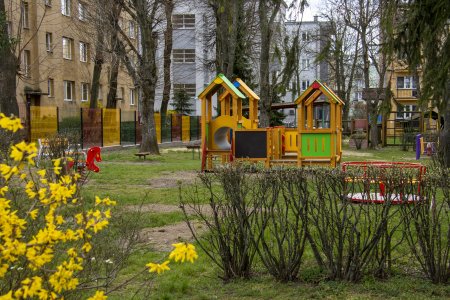 Przedszkolny plac zabaw z kolorowymi urządzeniami. Za placem zabaw widać blok.