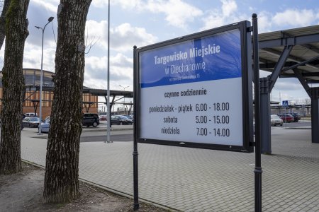 Tablica informacyjna przy części targowiska na wolnym powietrzu, pod zadaszeniami. Na tablicy znajdują się godziny, w których targowisko jest czynne.