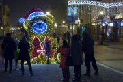 Zdjęcie po zmierzchu. Grupa ludzi fotografuje małą dziewczynkę, która ustawiła się do zdjęcia przy ozdobie świątecznej na deptaku - ogromnym misiu, który połyskuje setkami migoczących lampek