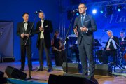 Na scenie Krzysztof Kosiński mówi do mikrofonu. Obok stoją: prowadzący koncert Jacek Borkowski i tenor Bogusław Morka. Z tyłu widać muzyków kameralnej orkiestry