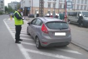 Strażnik miejski stoi obok samochodu. Wypisuje mandat kierowcy, który zaparkował auto na oznaczonej białymi liniami, wyłączonej z użytkowania części jezdni. W oddali budynek poczty.