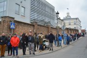 Rząd ludzi ustawionych na chodniku wzdłuż ulicy Wodnej. Za ich plecami ratusz z nowoczesną dobudowaną częścią otoczone zabytkowym ceglanym murem